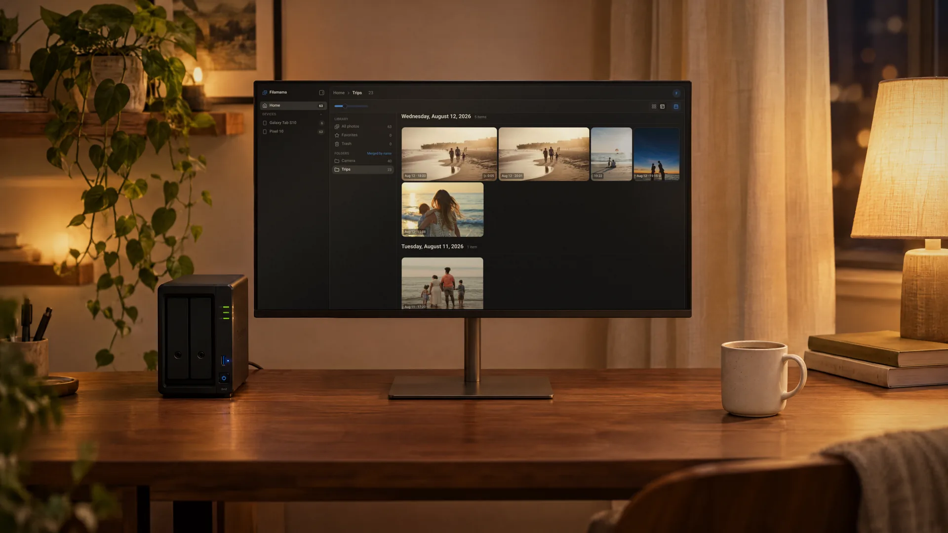A black two-bay Synology-style NAS next to a monitor on a walnut desk at night, the monitor showing the Filamama Trips folder in mosaic view