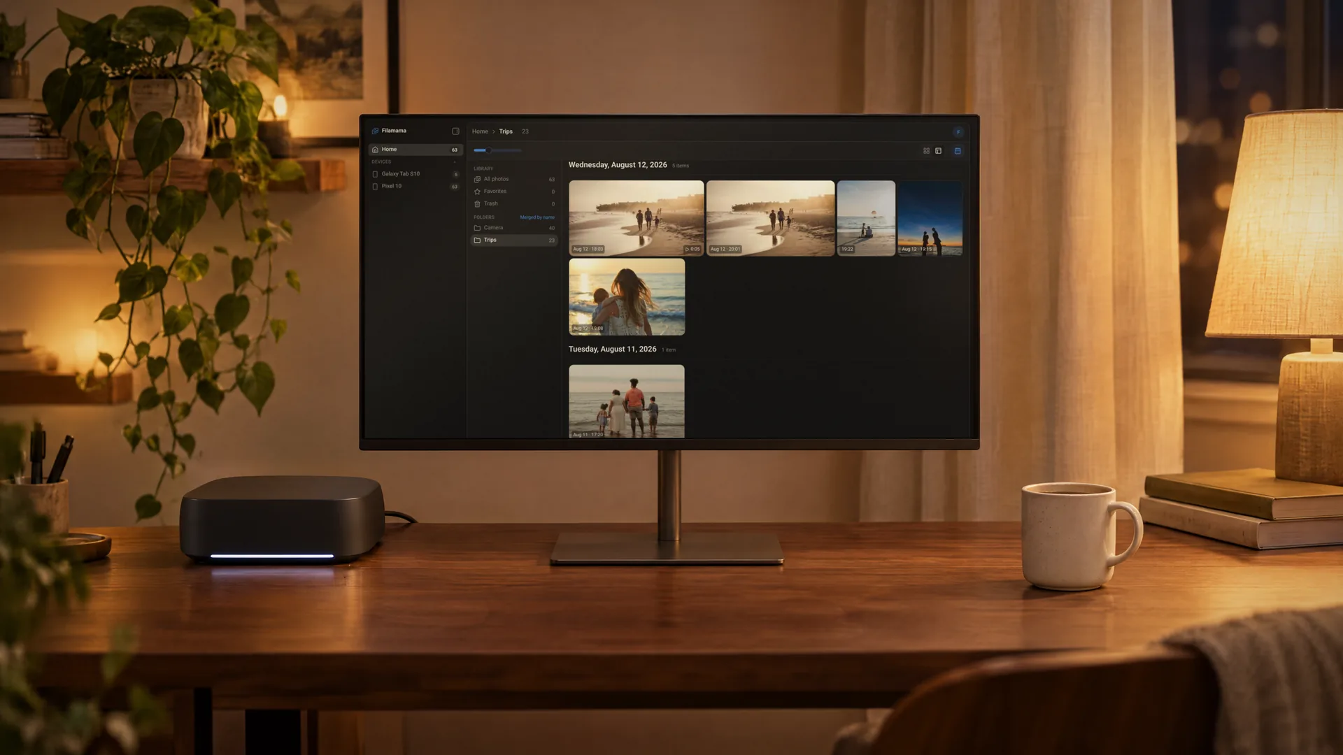 A rounded Umbrel-style home server next to a monitor on a walnut desk at night, the monitor showing the Filamama Trips folder in mosaic view